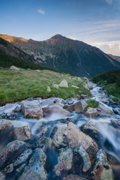 View of a stream cascading over rocks, flanked by verdant grass leading to a majestic mountain under a blue sky in Pribylina, Zilina Region, Slovakia.
