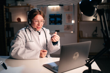 Female doctor in uniform wear headset making money gesture use laptop computer talking on video call with patient in hospital during night shift. Consulting, telemedicine and therapy