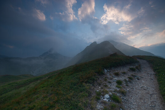 View of a rugged path winding along a verdant ridge under a dramatic sky, with peaks shrouded in mist, creating an ethereal landscape, Jamnik, Zilina Region, Slovakia.