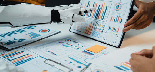 A human and a robot collaborate on data analysis, surrounded by various graphs and charts. Highlighting modern technology's role in business decision-making processes.Noogenesis