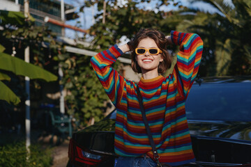 Woman in her mid 20s wearing a colorful striped sweater and yellow sunglasses, smiling and leaning against a black car in a tropical outdoor setting, casual outfit with relaxed travel vibe.