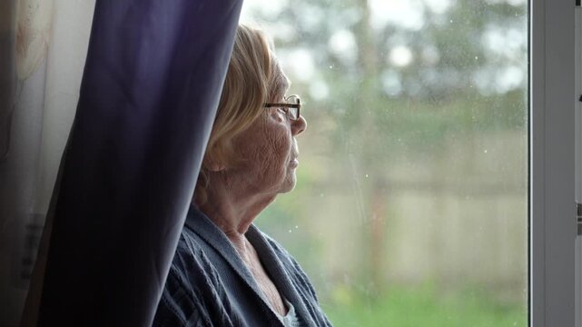 Senior lady gazes pensively outside. Aged woman peers through window glass thoughtfully. Elderly caucasian female contemplatively observes weather outside from rain spotted window. Lonely people