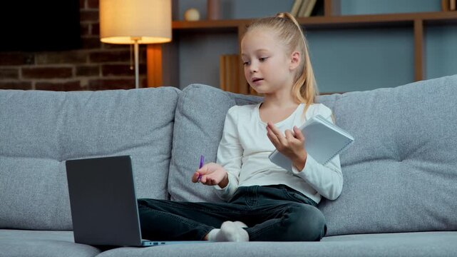 Happy Caucasian daughter girl kid little child writing studying with laptop on couch sofa e-learning remote home schooling write school homework listening online video call course lesson isolation
