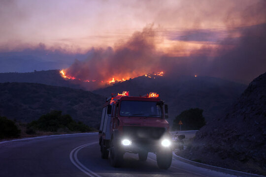 Firefighting truck with flashing lights drives on a road towards a large wildfire at dusk