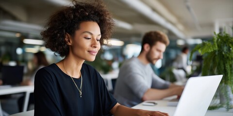 African female adult typing on laptop in modern office with caucasian male colleague in background