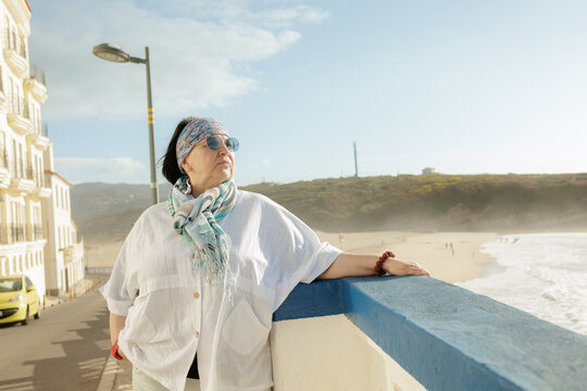 A cheerful mature woman in casual attire strolls happily along a sunny seaside promenade with waves crashing nearby. Stock photo - Powered by Adobe