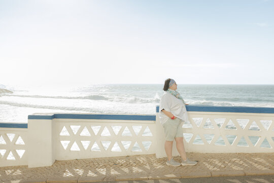 A cheerful mature woman in casual attire strolls happily along a sunny seaside promenade with waves crashing nearby. Stock photo - Powered by Adobe