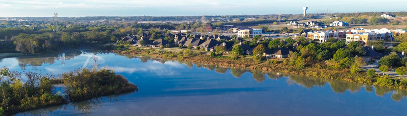 Obraz premium Panorama aerial tranquil lakeside area in Lantana, Texas, tree-lined streets, dark-roof homes, reflective water body bordered by seasonal foliage, suburban infrastructure, commercial buildings