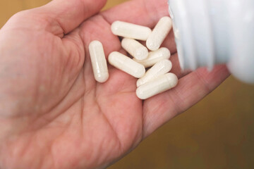 Close up of adult hand holding several white capsules poured from plastic bottle, daily supplement intake, health care routine, pharmacy, nutrition and wellness industry visuals