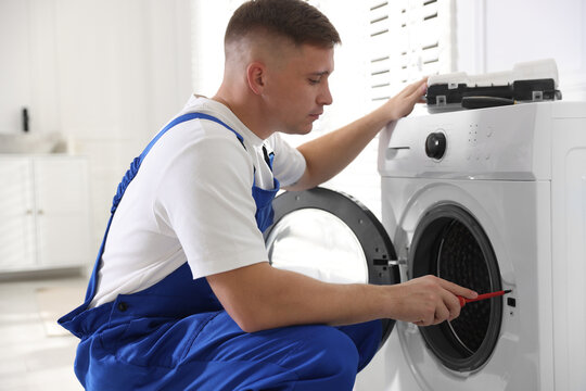 Repairman in uniform fixing broken washing machine with screwdriver at home
