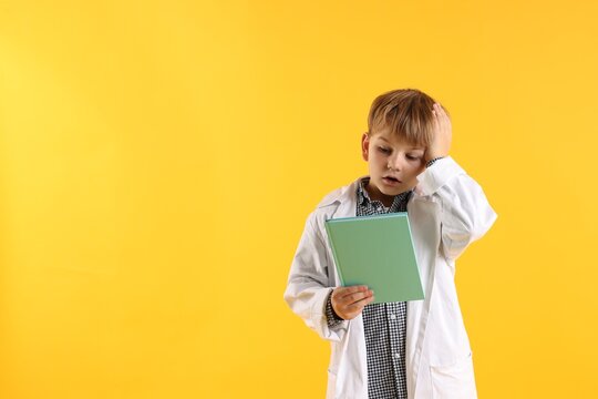 Emotional little boy in laboratory coat with book on yellow background, space for text. Child and science - Powered by Adobe