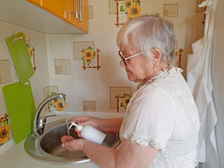 An elderly woman washes her hands with soap under running water from a faucet in a kitchen sink.