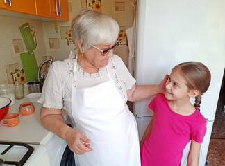 A grandmother with glasses and a white apron in the kitchen with her granddaughter.