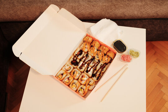 An open delivery box with assorted fried sushi rolls, soy sauce, ginger, wasabi, and chopsticks on a white table. A cozy interior setting highlights modern takeaway dining and Asian food culture