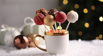 Tasty cake pops with sprinkles in cup on artificial snow against blurred lights, closeup. Christmas...