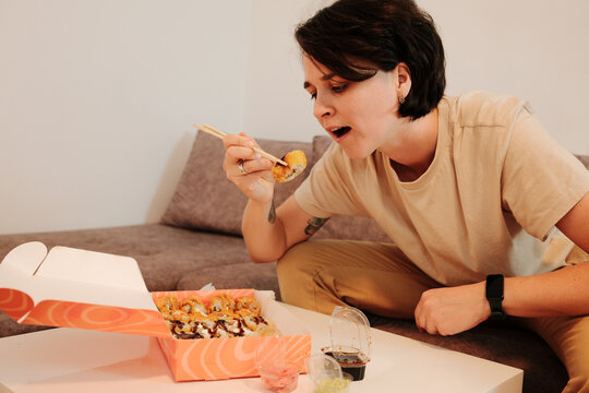 A woman sits on a sofa and eats fried sushi with chopsticks from a delivery box. The scene highlights casual dining, comfort, and modern home lifestyle with Asian food