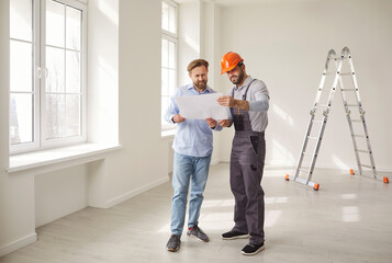 Construction team of two men standing in empty room and looking at blueprint together. Bearded...