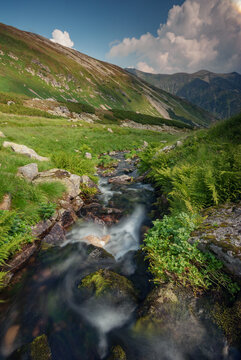 View of a creek cascading through a vibrant green meadow with mountains rising to meet a partly cloudy sky, Jamnik, Zilinsky region, Slovakia.