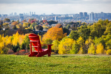 Fototapeta premium Red wooden chair on a grassy hill overlooking autumn trees and the distant city skyline of Toronto on a bright fall day