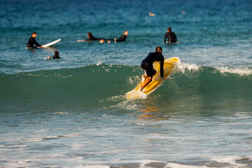 A male surfer wearing a wetsuit and paddling a yellow longboard on his knees through a wave with...