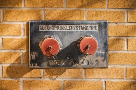 Fire department connection with dual standpipe and auto-sprinkler caps mounted on a brick wall in bright sunlight