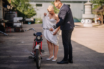 Bride in white dress adjusts her outfit while standing beside a vintage bicycle, with a groom in a suit assisting her in a rustic outdoor setting