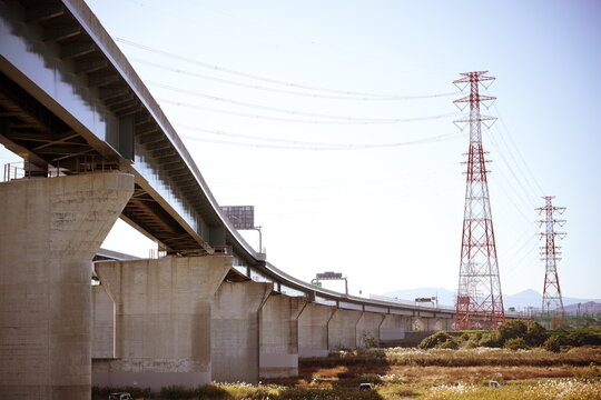 青空の下で高くそびえ立つ送電鉄塔と川岸の草地に沿ってカーブを描く高架道路の巨大なコンクリートの橋脚を捉えた景観