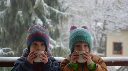 Two children in winter hats drinking a hot drink outdoors while it snows