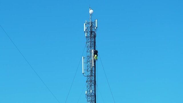 Repairmen install new cable and equipment. Workers repair cell tower. Two workmen on cell tower. Cell tower on blue background. Workmen, maintenance staff at work
