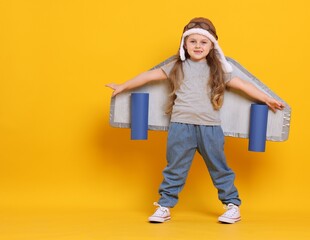 Little girl with cardboard plane wings and hat playing pilot on orange background