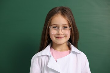 Smiling little girl in laboratory coat and glasses near green chalkboard. Child and science