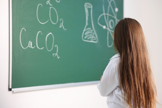 Little girl in laboratory coat near green chalkboard with formulas indoors, back view with space for text. Child and science
