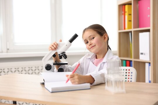 Cute little girl with pen, book and microscope at desk indoors. Child and science