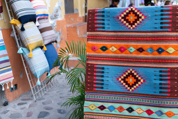 A colorful woven rug with intricate geometric patterns displayed alongside decorative cushions in an outdoor market setting.