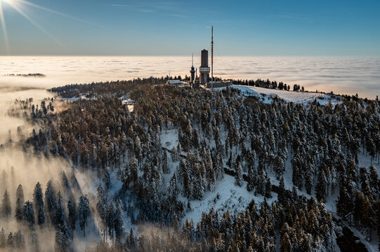 Aerial view of the Grosser Feldberg peak adorned with snow-laden trees and a prominent tower piercing through the clouds, Schmitten im Taunus, Hessen, Germany.