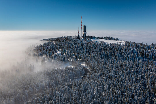 Aerial view of snow-covered Grosser Feldberg mountain peak and tower piercing through a sea of clouds under a clear blue sky, Schmitten im Taunus, Hessen, Germany.
