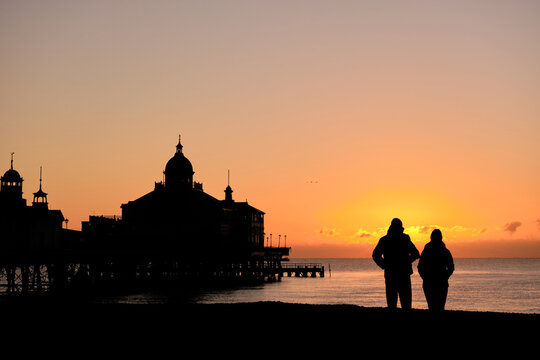 Silhouette of a couple standing on a beach watching the sunrise with a domed pier to their left.