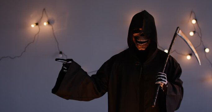 Man dressed black as grim reaper of death standing with scythe, pointing skeletal finger to side at something and nodding in Halloween festival, on dark illuminated background. Halloween party concept