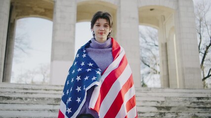 Young woman standing on steps wrapped in American flag during a sunny outdoor event in a park - Powered by Adobe