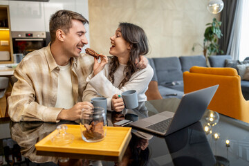 Couple enjoying coffee and cookies while working on laptop in cozy modern living room