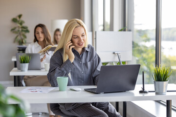 Obraz premium Woman working on a laptop and talking on the phone in a modern office setting with plants and natural light
