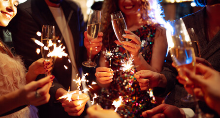 Hands hold sparklers, creating bright sparks in a lively setting. Friends celebrate together as...