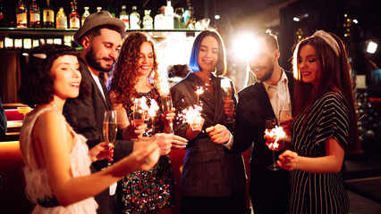 Friends gather at a vibrant bar to celebrate New Year's Eve. They enjoy drinks while holding sparklers, surrounded by a festive atmosphere and sparkling decorations.