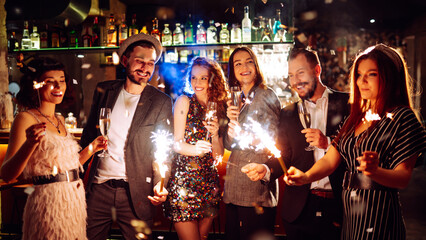 Friends gather at a vibrant bar to celebrate New Year's Eve. They enjoy drinks while holding sparklers, surrounded by a festive atmosphere and sparkling decorations.