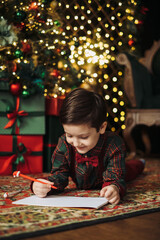 A child in a plaid shirt and bow tie writes a letter to Santa under the twinkling lights of a...