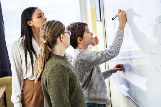 A few students are solving math problems on the whiteboard while the teacher watches from the side. The moment feels focused and active as they work through the task together. - Powered by Adobe