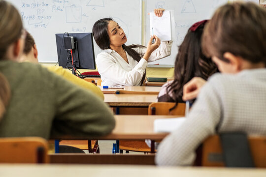 A small group of students is in their elementary classroom while a mixed-race teacher guides them through a math and geometry lesson. She is asking questions and helping them stay focused as they work