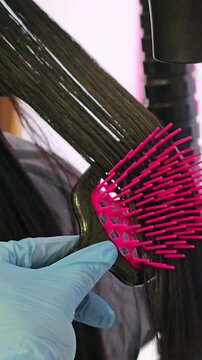 Hair stylist blow-drying and styling a woman's long hair in a beauty salon. Vertical close-up video of stylist using hair dryer and comb to dry and shape dark hair in beauty salon