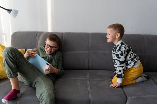 Two brothers laughing on grey sofa with older boy holding tablet while younger sibling watches happily at home. Concept of siblings playful interaction, shared humor, joyful childhood moments together