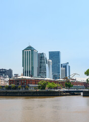 Obraz premium Vista vertical de Puerto Madero mostrando el contraste entre los antiguos docks de ladrillo rojo, una grúa portuaria y los modernos rascacielos corporativos frente al río bajo un cielo azul.